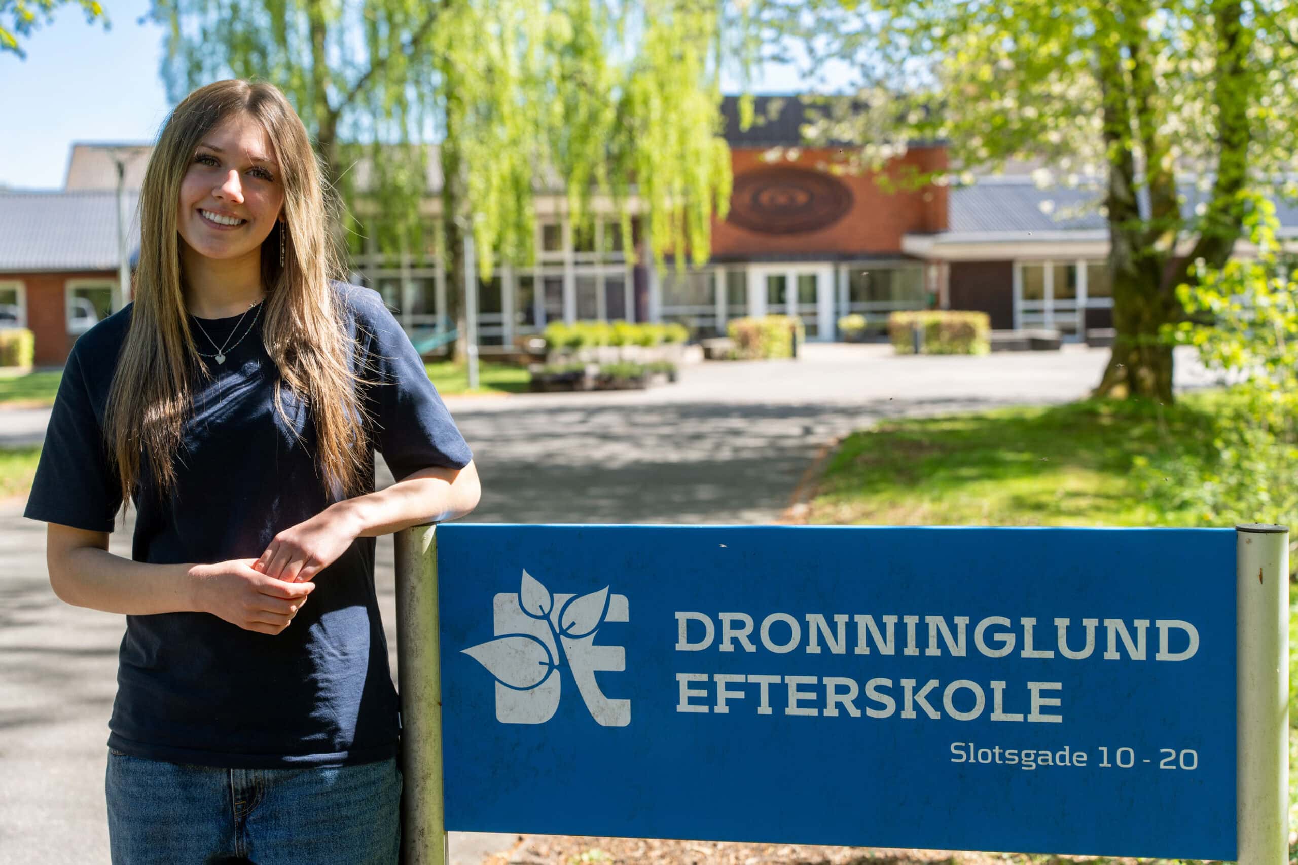 Dronninglund Efterskole - Elev foran skolens skilt med grønne træer i baggrunden.